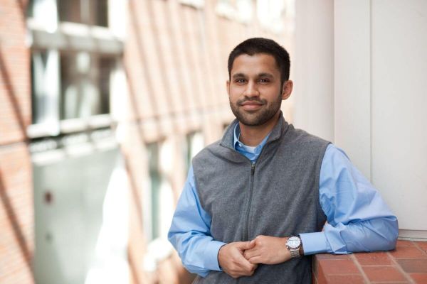 A University of Rochester student in a vest and blue shirt leans against a brick wall, looking relaxed and contemplative.