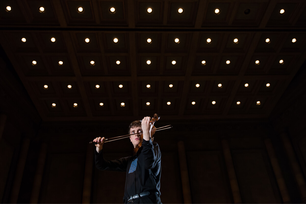 A University of Rochester Eastman School of Music student holds a violin beneath an ornate ceiling.