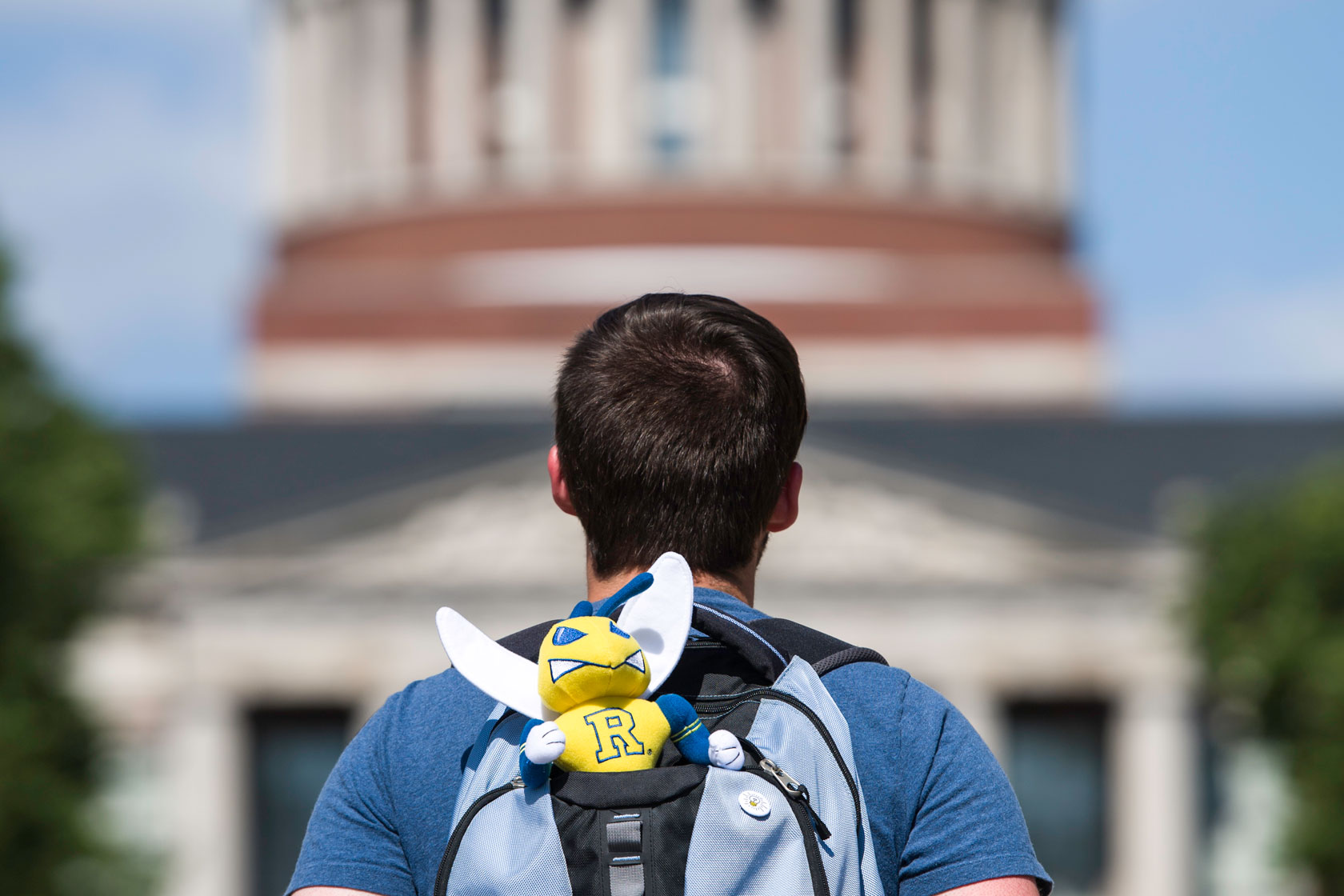 A person with short brown hair is seen from behind, wearing a blue shirt and carrying a light blue and gray backpack. Attached to the backpack is a small plush mascot with a yellow face, blue wings, and a letter "R" on its chest. A dome-shaped building is blurred in the background.