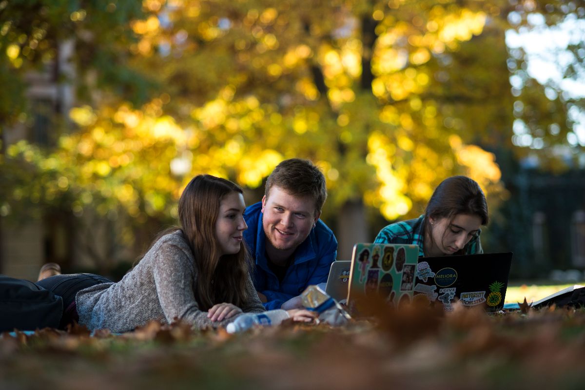 Three URochester students sitting on the grass, focused on their laptops, enjoying a sunny day outdoors.