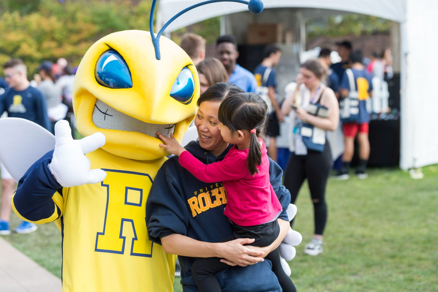 A woman and child embrace a cheerful mascot at a University of Rochester event, showcasing joy and community spirit.