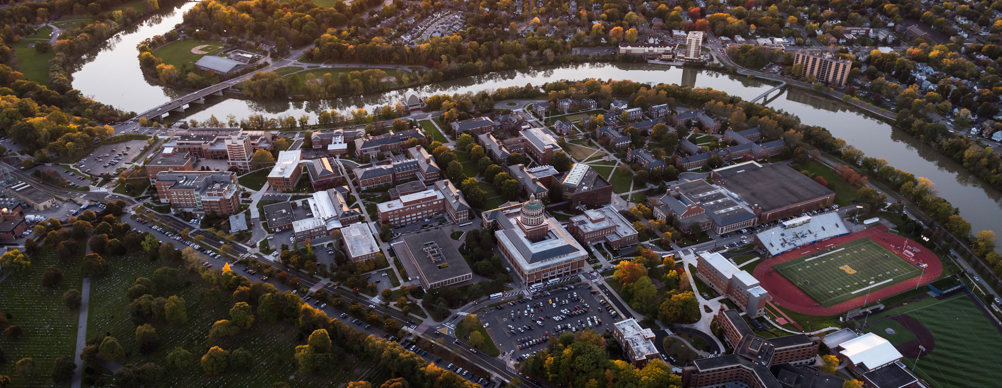 Aerial view of the University of Rochester campus, featuring green spaces and a river winding through the landscape.