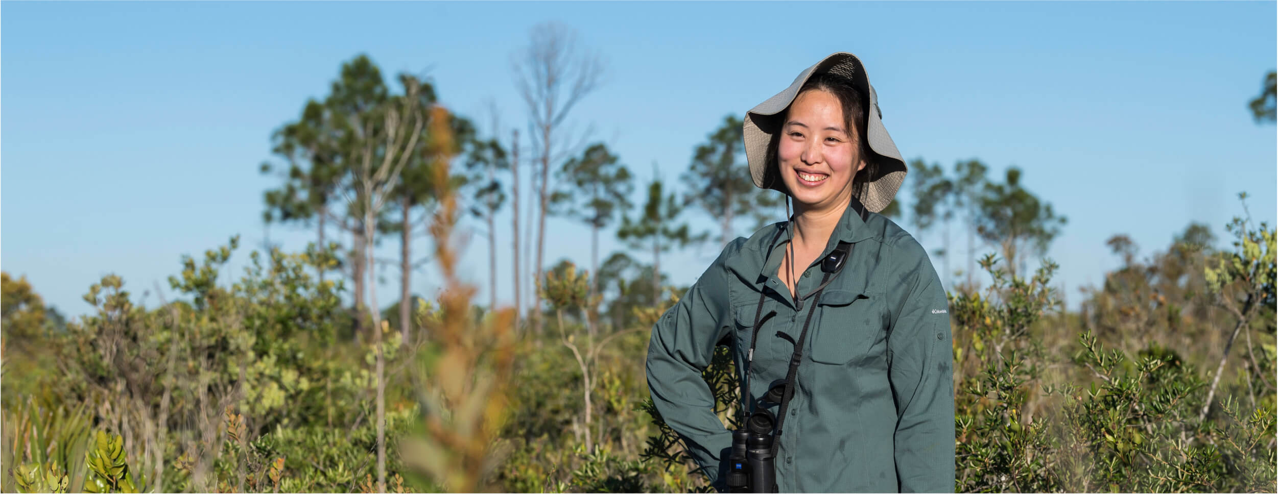 A woman in a hat and green shirt stands in a field, representing a University of Rochester research faculty member.