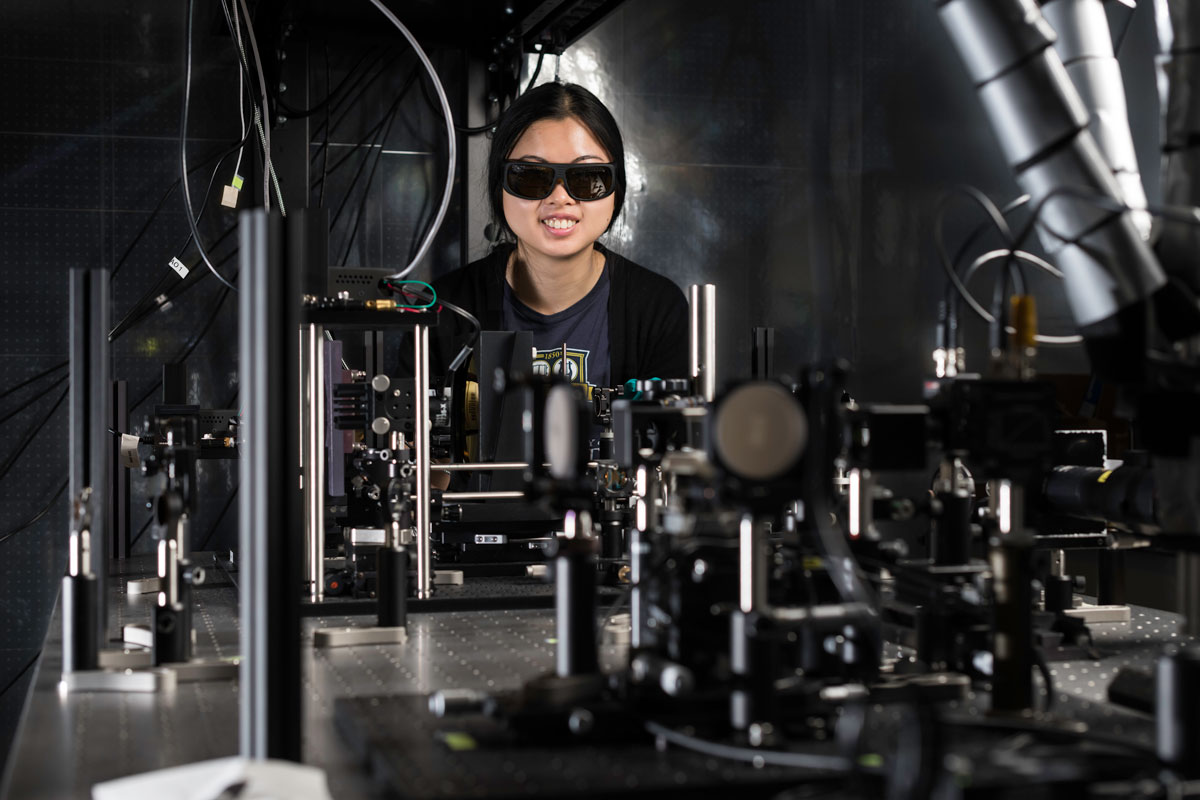 A woman wearing sunglasses operates a machine in a University of Rochester lab setting.