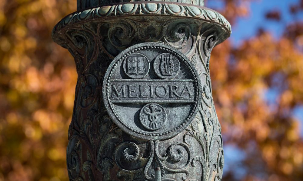 A flagpole bass  featuring a sign that says meliora situated on the University of Rochester grounds.