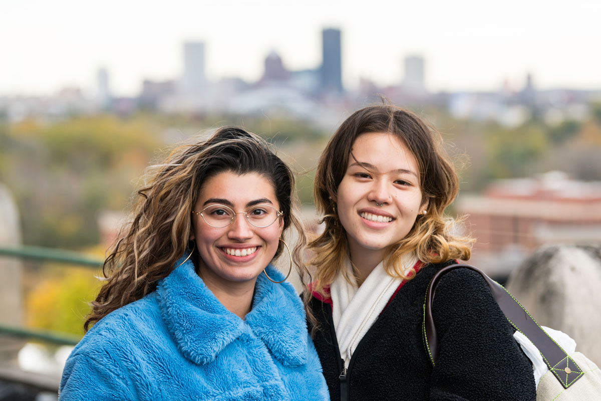 Two women smiling at the camera on the University of Rochester campus, with the city skyline visible in the background.