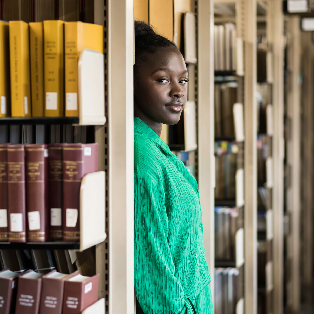 A University of Rochester student stands confidently in front of a bookshelf filled with various books.