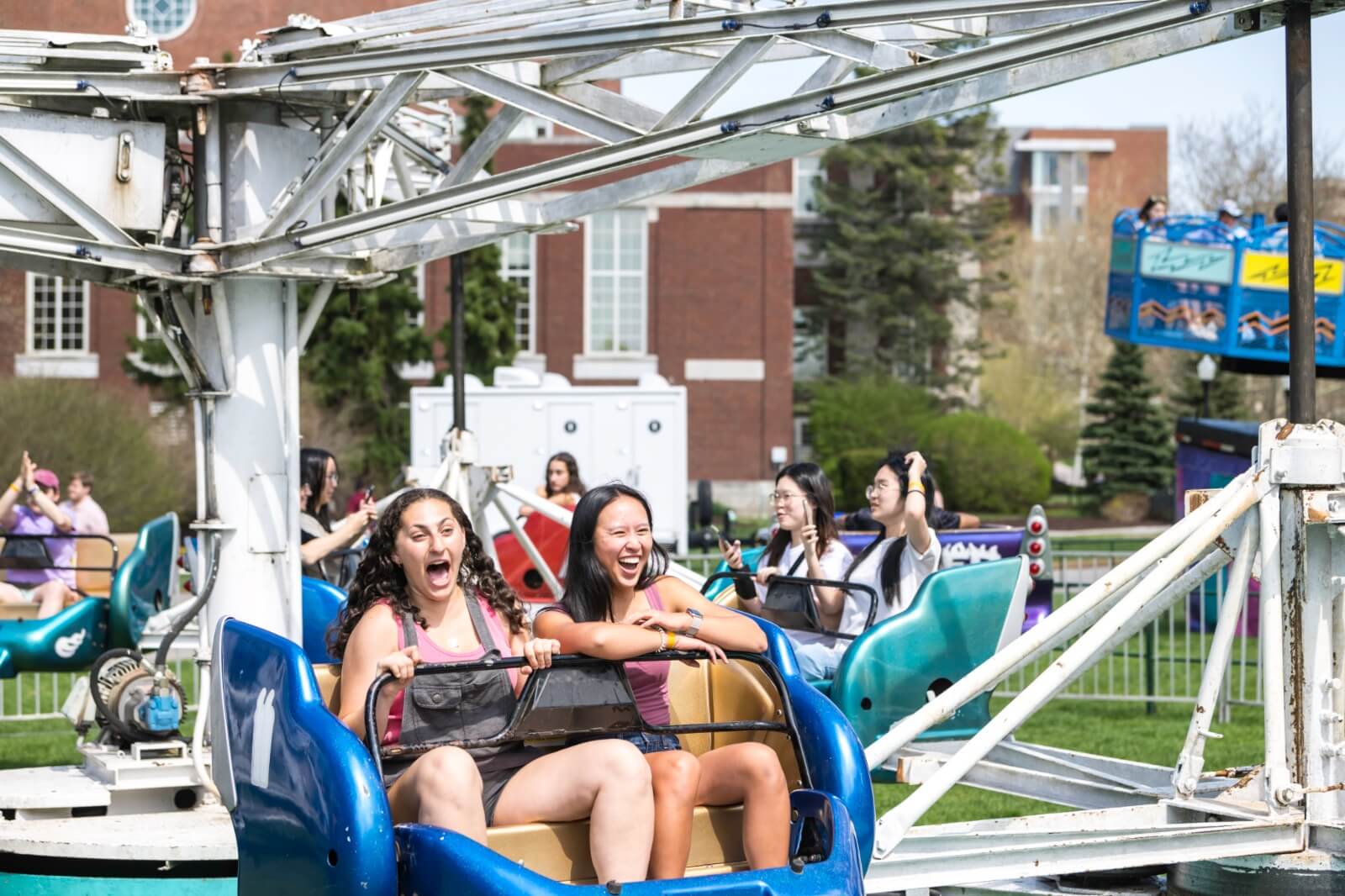 Two young women are laughing and riding a spinning amusement park ride outdoors. Other people are also enjoying the ride in the background. The scene is sunny and cheerful with buildings and trees visible in the background.