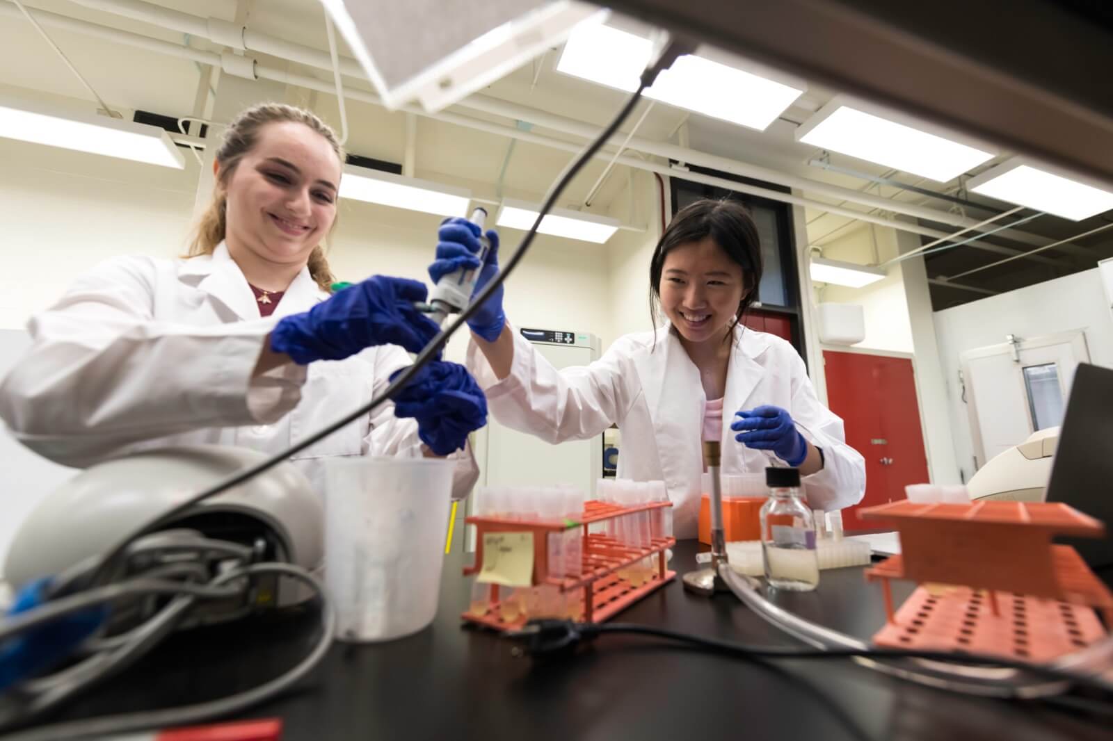 Two scientists in lab coats and gloves work in a laboratory. One uses a pipette while the other smiles and observes. Test tubes, beakers, and lab equipment are on the black workbench. The environment is bright with institutional lighting and equipment around them.