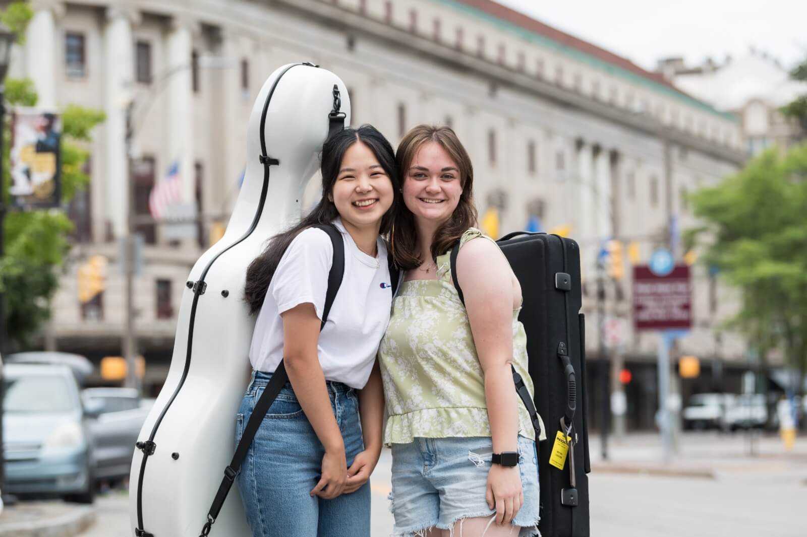 Two smiling young women stand close together, each carrying instrument cases on their backs. They are outdoors in an urban area with blurred buildings, vehicles, and street signs in the background. One wears a white shirt and jeans, the other a green top and shorts.