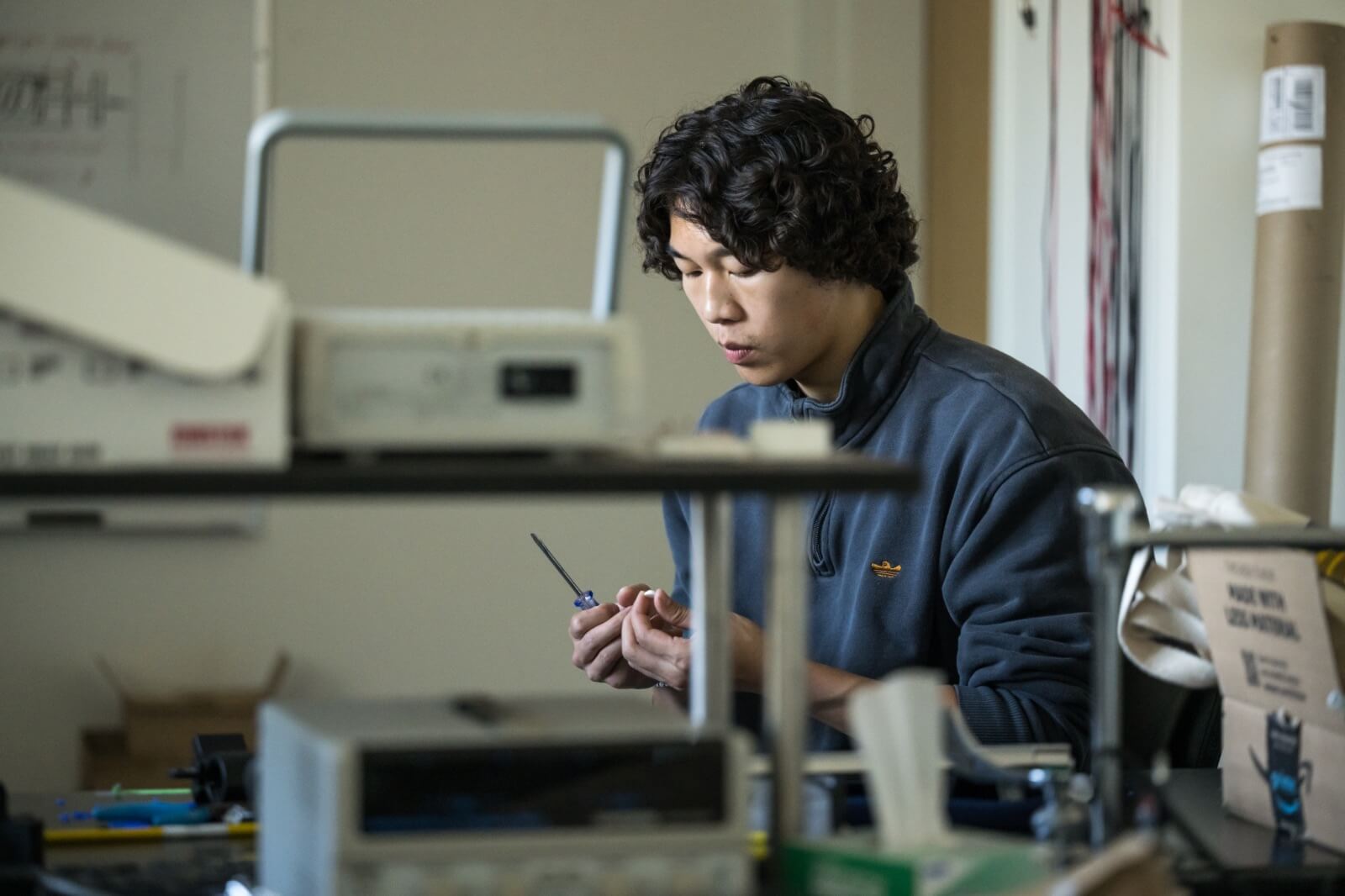 A person with curly hair, wearing a dark sweatshirt, focuses intently on working with a small tool in a cluttered workshop. The room is filled with various equipment and materials.