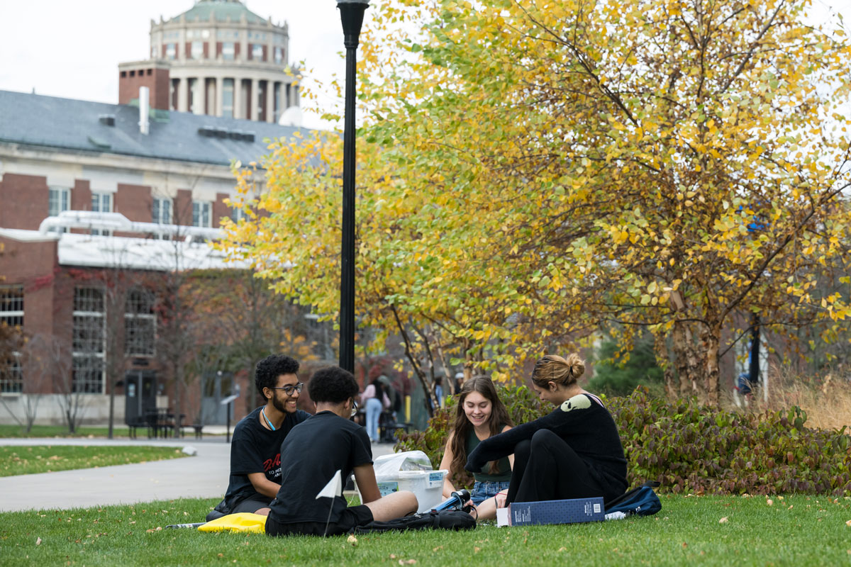 Students relax on the grass in front of the University of Rochester library, enjoying a sunny day outdoors.