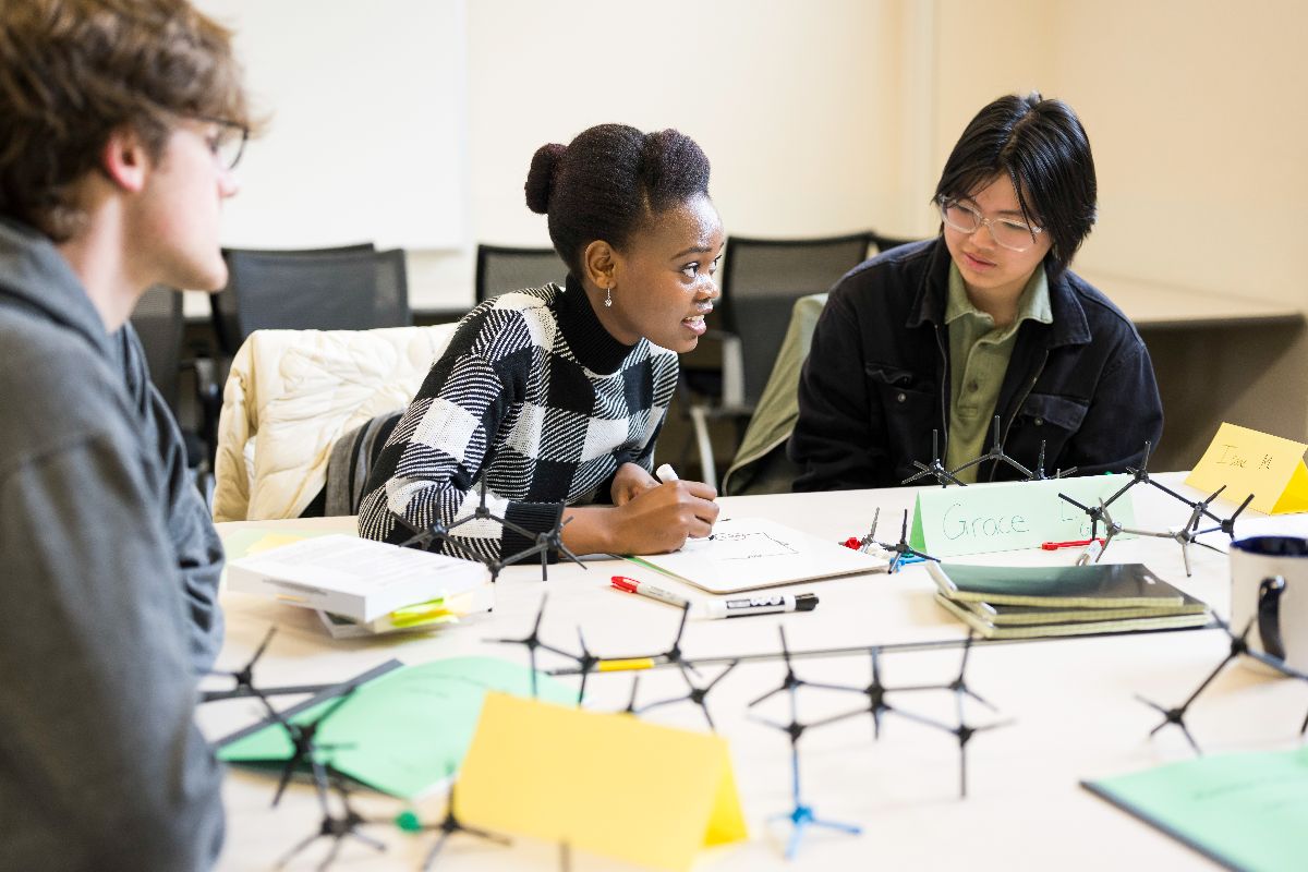 Three URochester students collaborate at a table, surrounded by a notebook and pencils, engaged in study.