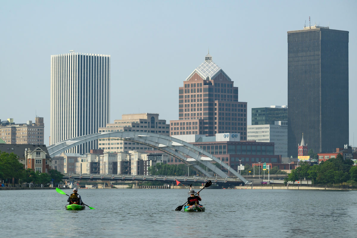 URochester students kayak on the Genesee River with a large city skyline in the background, showcasing Rochester, New York.