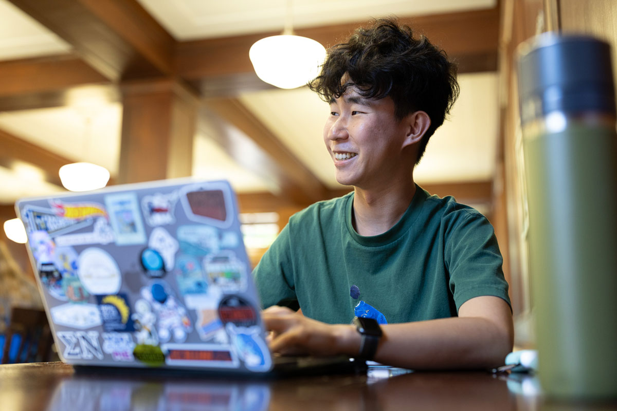A young University of Rochester student smiles while working on a laptop, engaged in his studies.