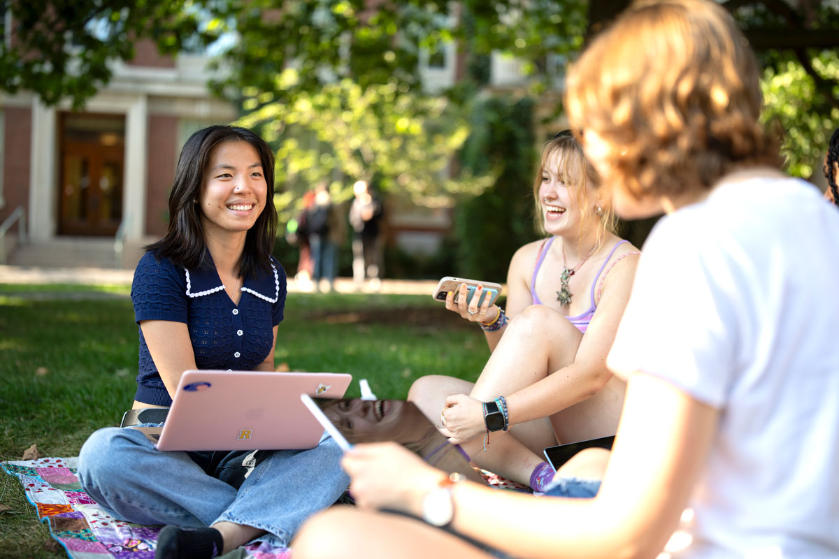 Three University of Rochester students sitting on the grass, working on laptops in a sunny outdoor setting..
