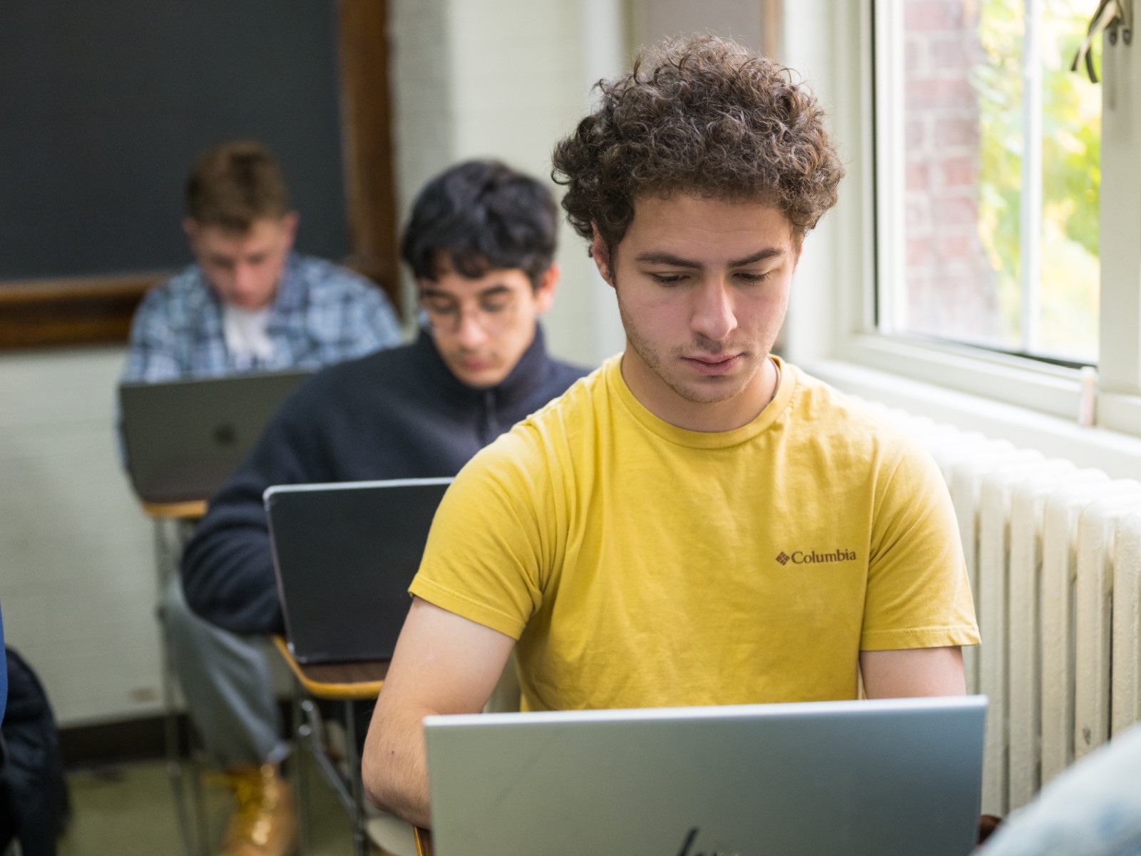 Three students sit at desks one in front of another in a classroom while looking at their laptops.