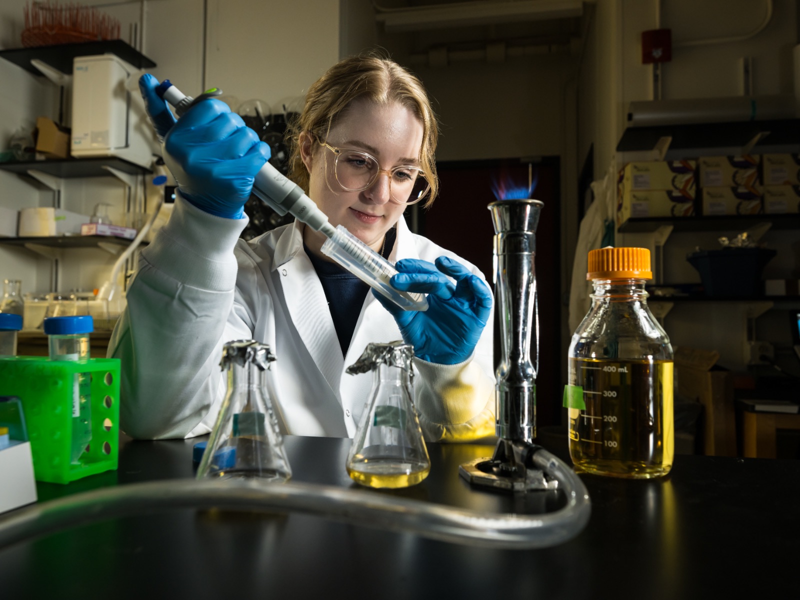 Student in labcoat sitting at desk uses equipment and test tube with various beakers in front of her.