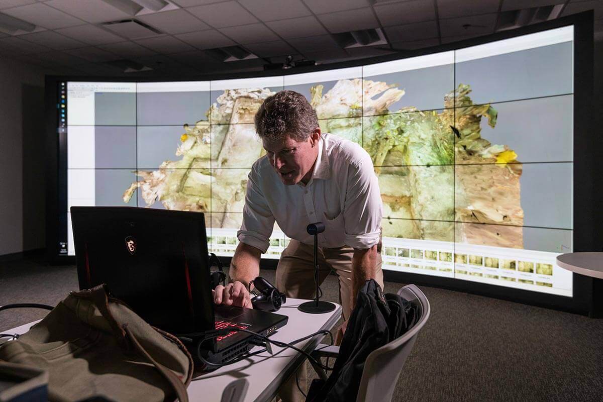 Man leaning over laptop with curved wall monitor display in background at University of Rochester.