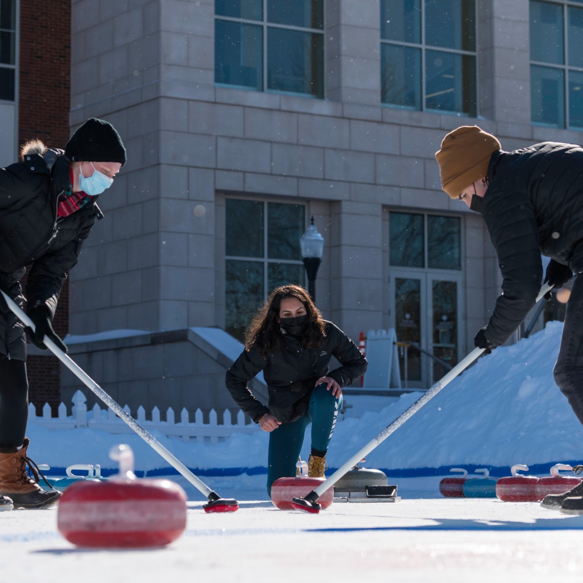 Three people wearing masks participate in curling in winter. One is on their knee, and two are sweeping