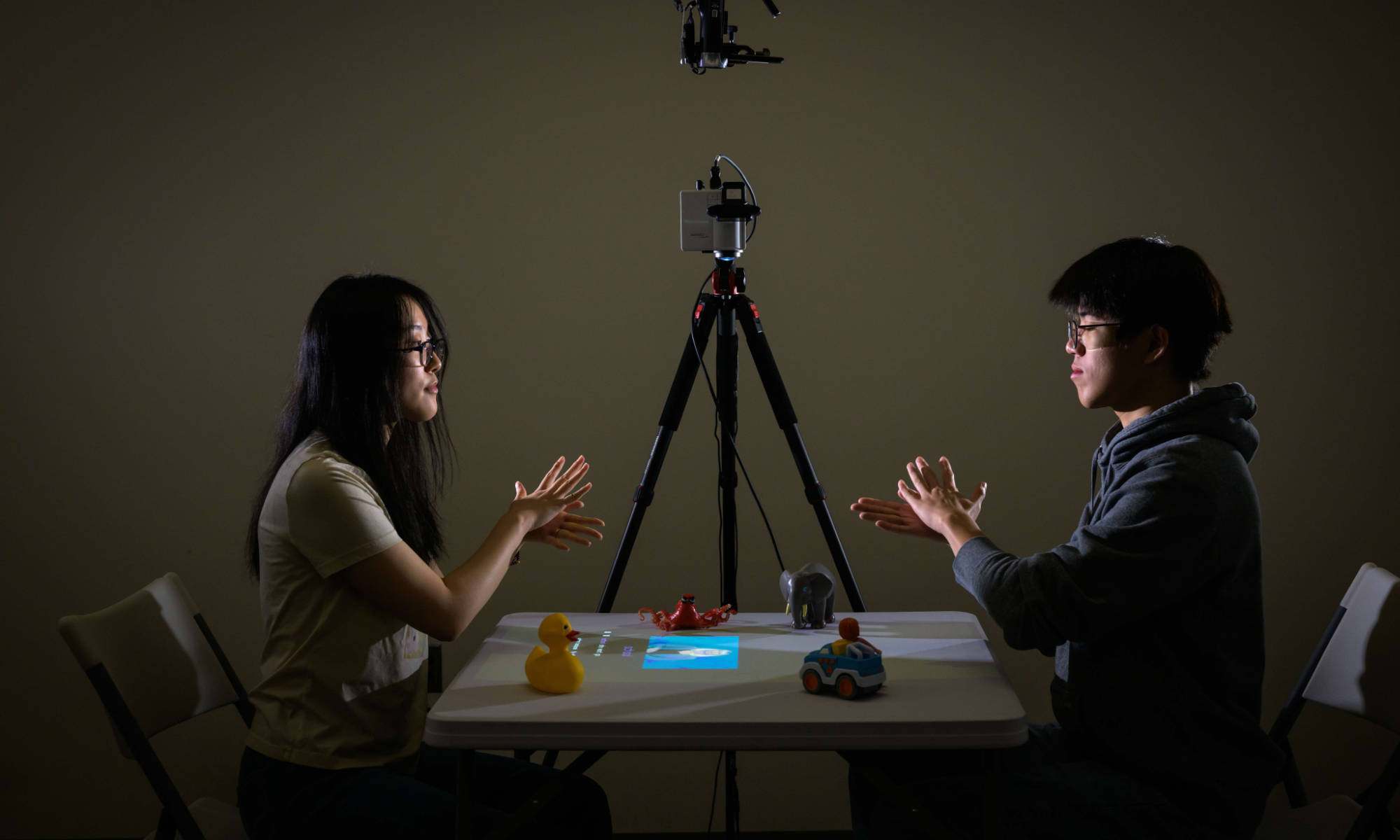 Two people sit across from a table using American Sign Language, artificial intelligence, and augmented reality to test a tabletop interactive play system for deaf and hard of hearing children.