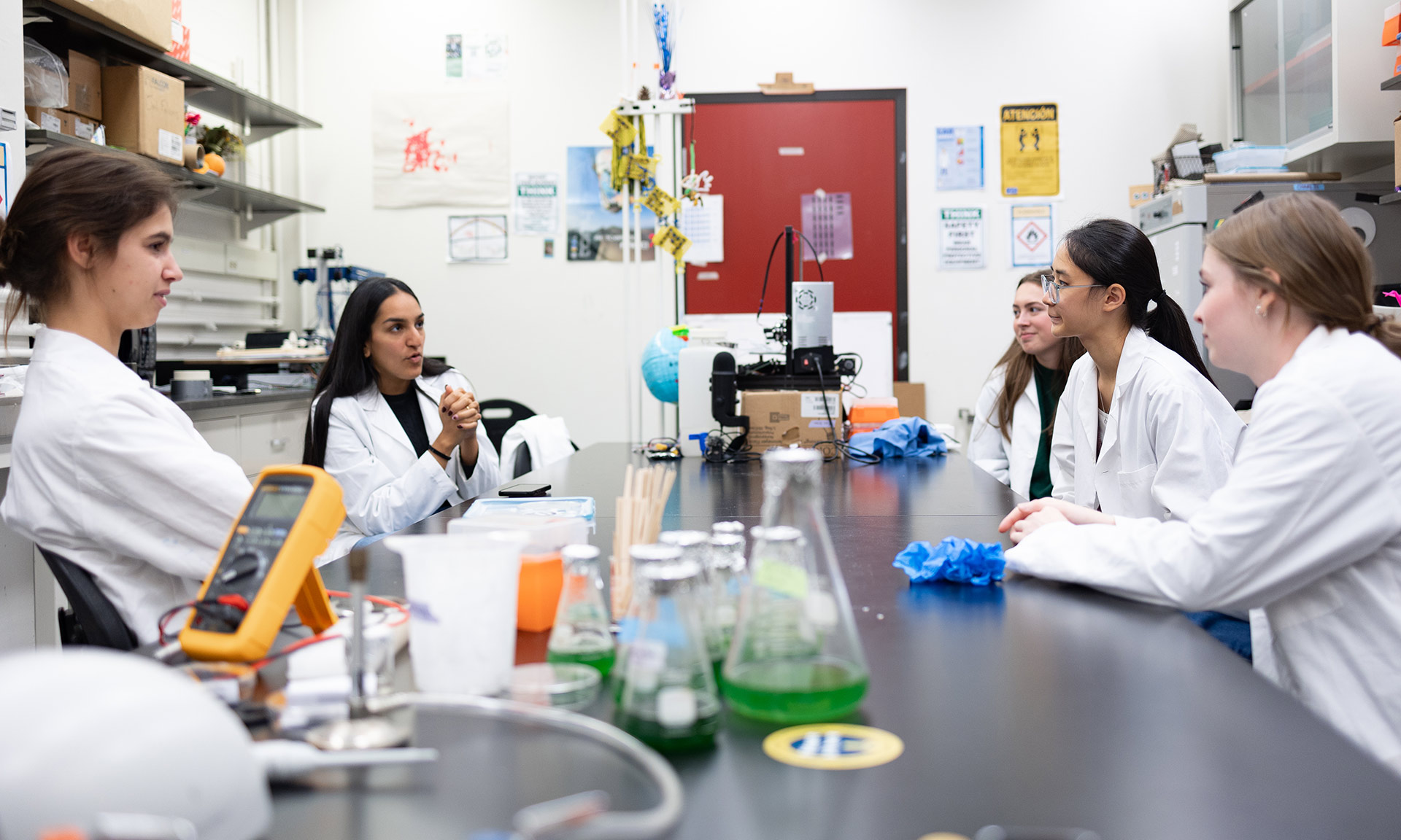 Group of five students who work on synthetic biology projects in white lab coats sitting around a table filled with beakers and others scientific equipment.
