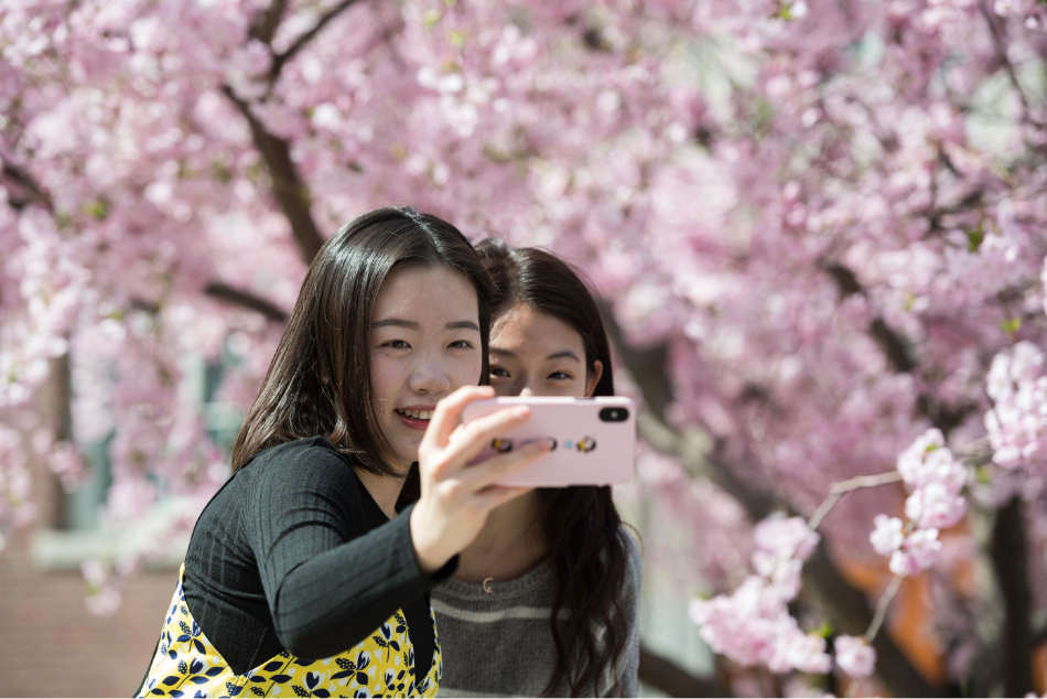  Two young women smiling while taking a selfie in front of a flowering tree at the University of Rochester.