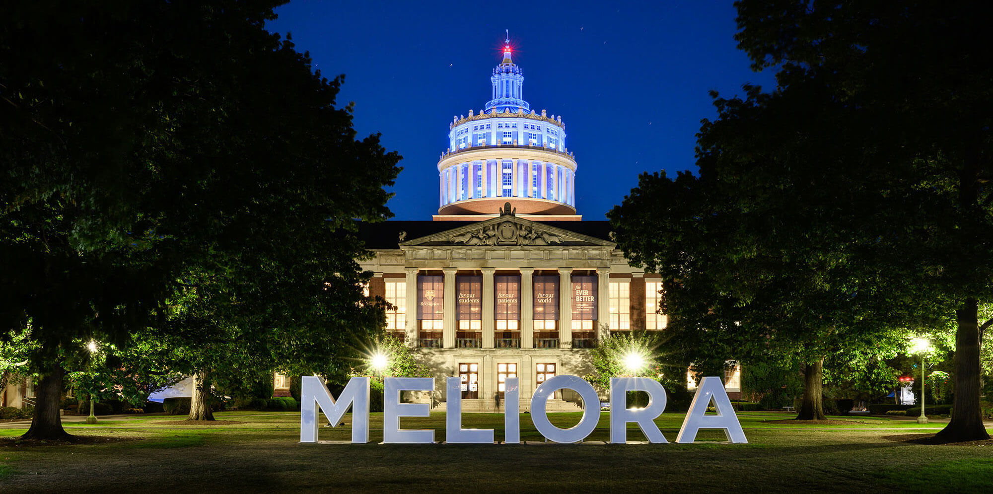 A large illuminated “MELIORA” sign sits on the lawn in front of Rush Rhees Library at the University of Rochester, with the dome lit in blue against the night sky.