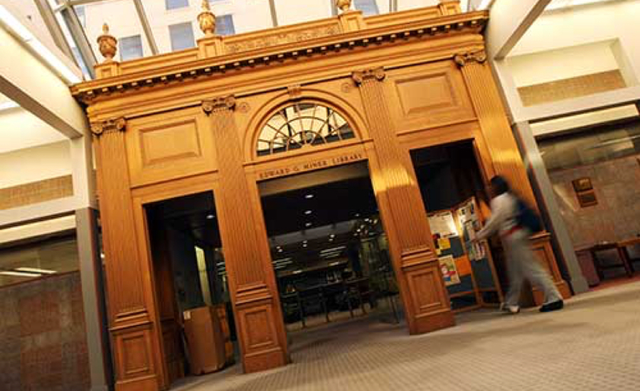 A person walks through the Edward G. Miner Library at the University of Rochester, passing a large wooden door.
