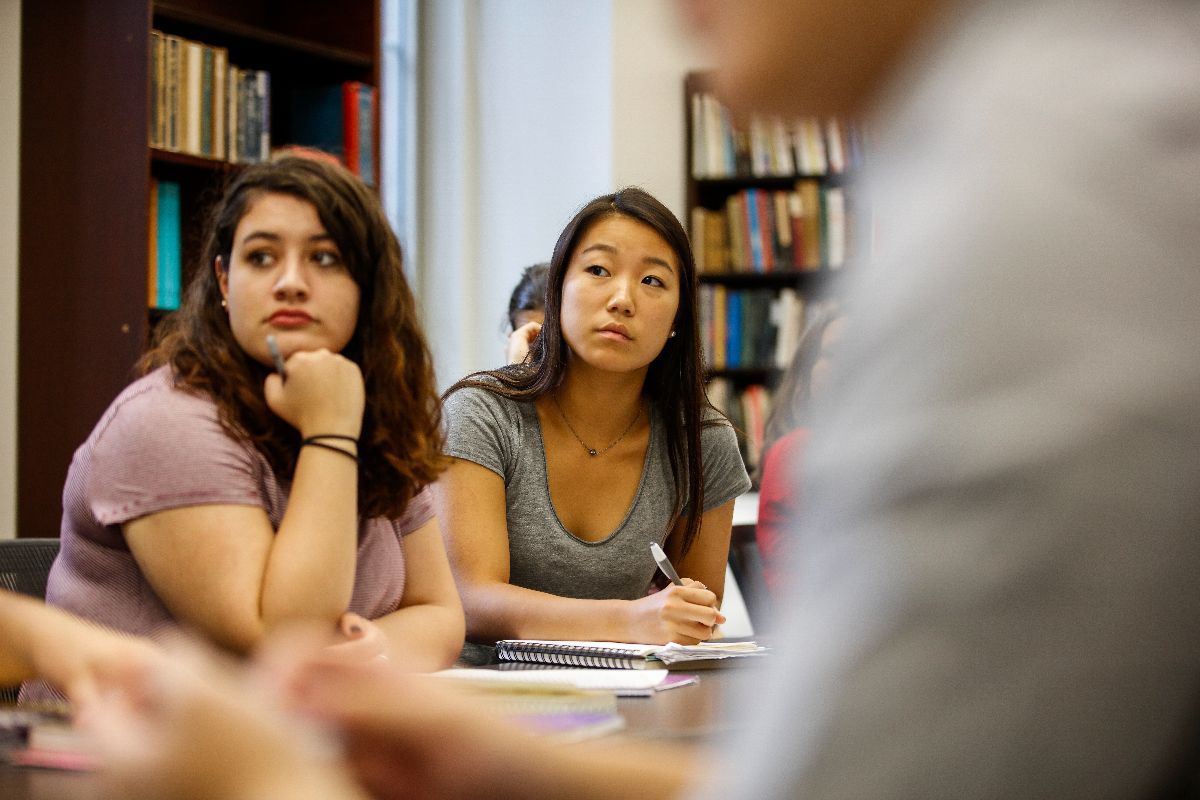 URochester students engaged in a classroom discussion, seated at desks with books and laptops open.
