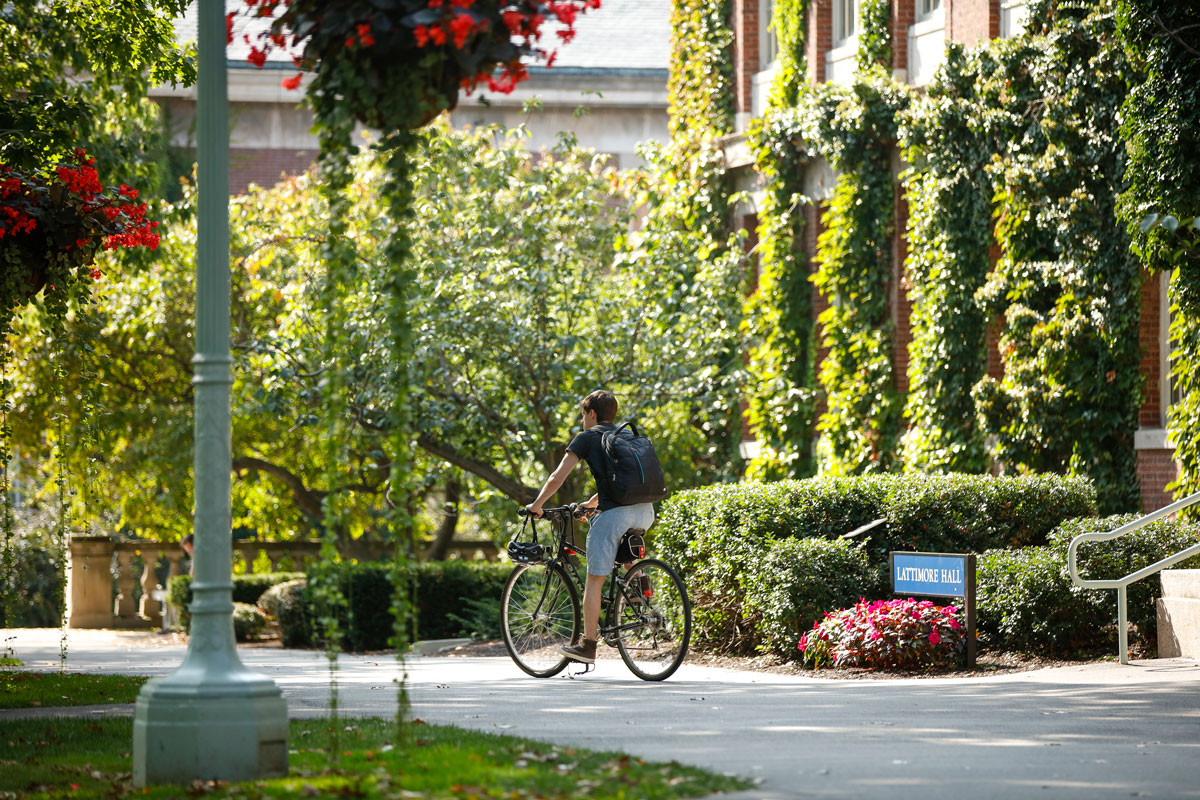 A man rides a bike on a sidewalk at the University of Rochester campus, surrounded by greenery.