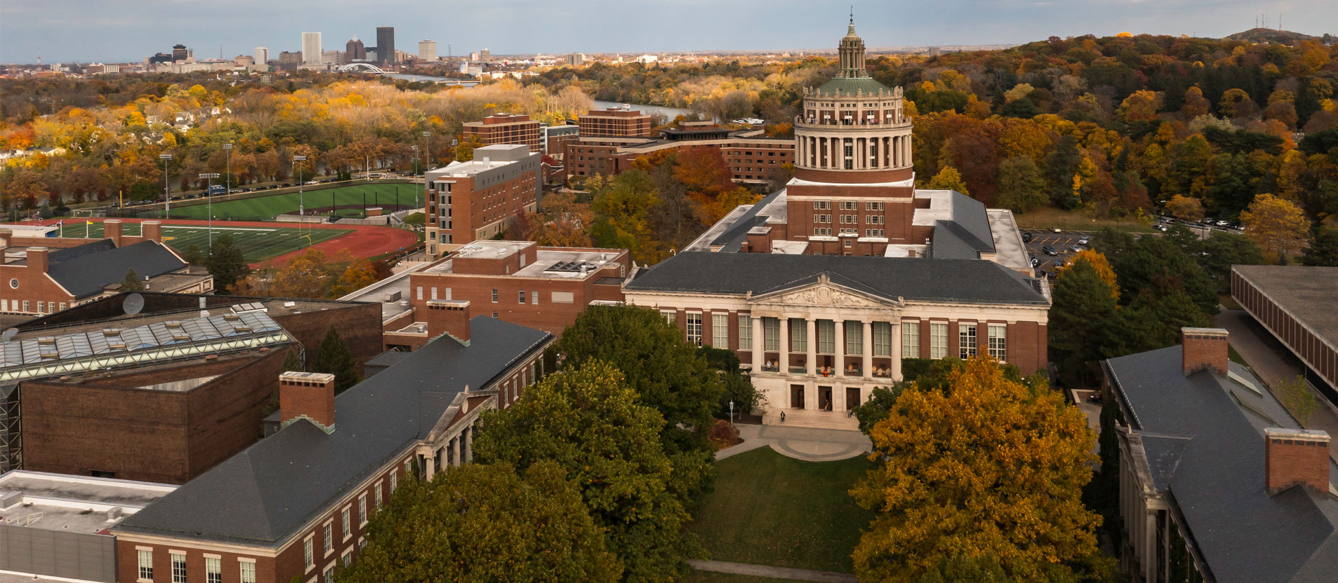 Aerial view of a university campus with a prominent dome-topped building amid fall foliage and a distant city skyline.