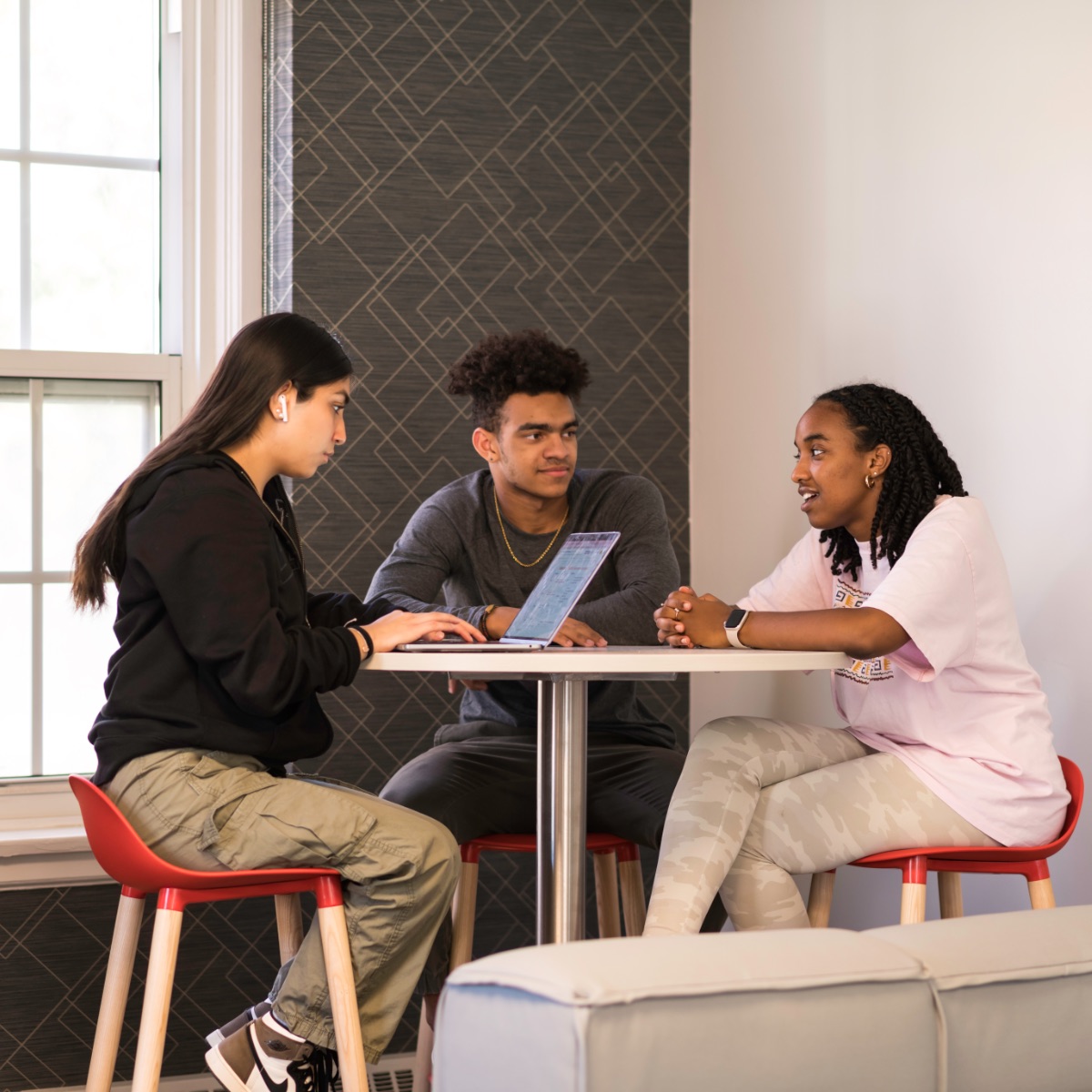 Three students sitting at a round desk. One looks at their laptop