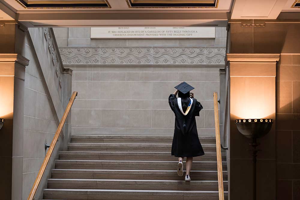 A University of Rochester student walks up stairs of Rush Rhees Library in cap and gown