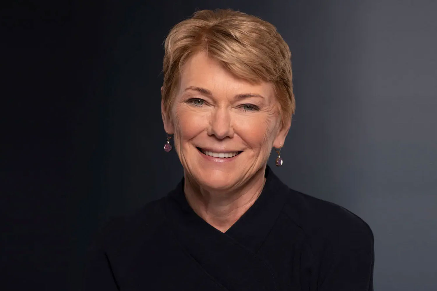 President Sarah C. Mangelsdorf, wearing a blue blazer and black shirt, stands in front of a bookshelf at the University of Rochester.